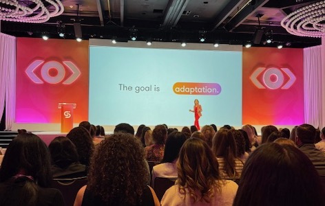 A speaker presenting on stage at a conference, with a slide displaying the text 'The goal is adaptation' in colorful letters, and an audience seated in front of them.